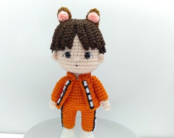 May include: A crocheted doll with brown hair, orange and white clothing, and brown ears. The doll is standing on a silver surface.
