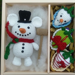 Może przedstawiać: Śniegowy ludzik wykonany na szydełku z czarnym kapeluszem i czerwoną szalikiem, prezent wykonany na szydełku w kolorze zielonym i czerwonym, niebieski śniegowy ludzik wykonany na szydełku oraz czerwono-biała truskawka wykonana na szydełku w pudełku prezentowym.