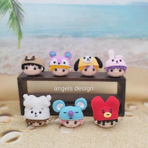 Puede incluir: Una colección de siete muñecos de ganchillo hechos a mano, cada uno con un diseño de sombrero único. Los muñecos se exhiben sobre una superficie de madera y arena, con una escena de playa al fondo. Los muñecos presentan varios colores, incluyendo rosa, azul, amarillo y rojo. El texto "angels design" es visible.