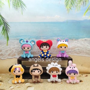 May include: Seven small crocheted dolls arranged on a dark wooden shelf. Each doll wears a different animal-themed hood, including a blue mouse, red bear, white rabbit, yellow dog, brown bear, white flower, and pink rabbit. The text angels design is visible.