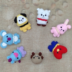 Könnte beinhalten: Eine Sammlung von sieben gehäkelten Plüschfiguren in verschiedenen Farben. Die Figuren umfassen einen gelben Hund, einen weißen Bären, ein rosa Kaninchen, einen blauen Koala, eine blau-violette Figur, eine braune Kuh und ein rotes Herz mit blauem Hut.