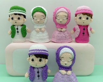 May include: Six crocheted dolls dressed in traditional Islamic clothing. The dolls are wearing green, white, pink, and purple clothing. The dolls are all standing on a white surface.