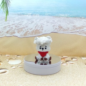 K&ouml;nnte beinhalten: Wei&szlig;es geh&auml;keltes Pl&uuml;schtier mit einem roten Bandana, das auf einem wei&szlig;en Sockel sitzt. Das Spielzeug hat schwarze Augen und eine schwarze Nase. Der Hintergrund ist eine Strandszene mit Sand und blauem Wasser.