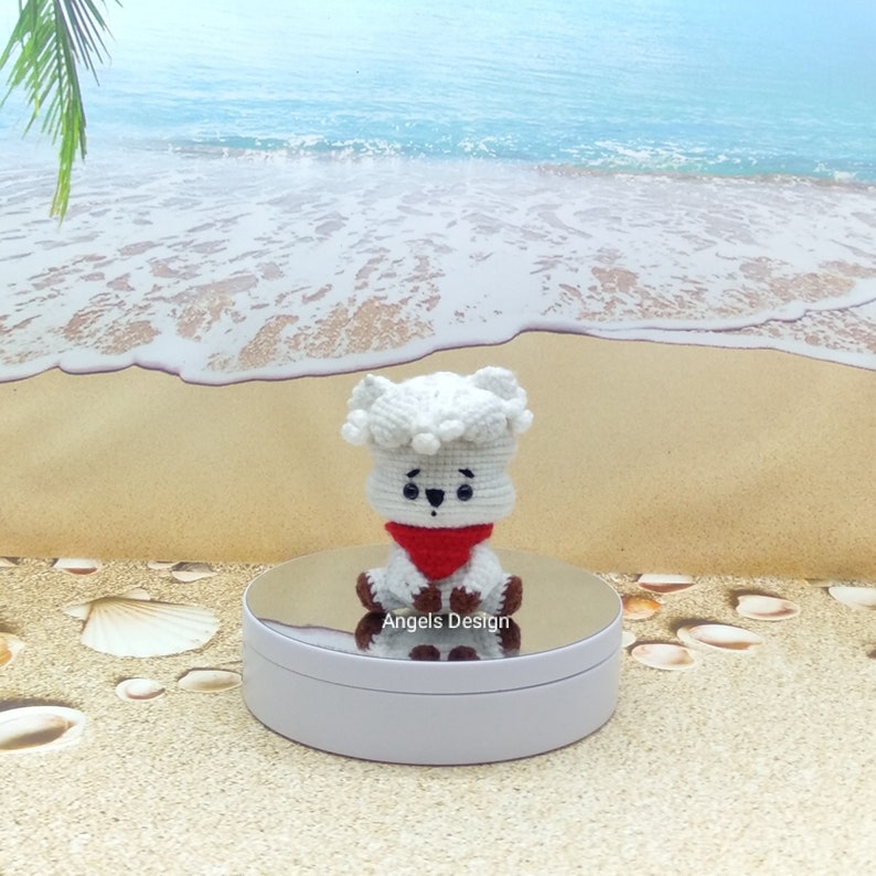 K&ouml;nnte beinhalten: Wei&szlig;es geh&auml;keltes Pl&uuml;schtier mit einem roten Bandana, das auf einem wei&szlig;en Sockel sitzt. Das Spielzeug hat schwarze Augen und eine schwarze Nase. Der Hintergrund ist eine Strandszene mit Sand und blauem Wasser.