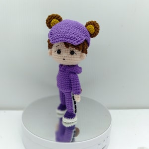 May include: A purple crocheted doll with brown hair, wearing a purple hat and purple pants with black stripes. The doll is standing on a white platform.