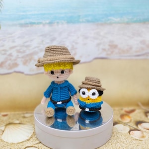 May include: A crocheted boy doll wearing a blue shirt, brown hat, and brown belt, sitting next to a crocheted yellow minion doll wearing a brown hat and blue overalls. Both dolls are sitting on a white circular platform.