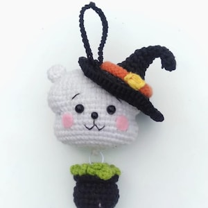 May include: A white crocheted ghost wearing a black witch hat with orange and yellow accents. The ghost is holding a black cauldron with a green rim.