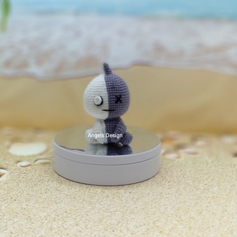 K&ouml;nnte beinhalten: Ein geh&auml;keltes Pl&uuml;schtier mit einem wei&szlig;en und violetten Design. Das Spielzeug hat ein einzelnes Auge und ein X f&uuml;r einen Mund. Das Spielzeug sitzt auf einem wei&szlig;en runden St&auml;nder mit einer verspiegelten Oberfl&auml;che. Der St&auml;nder steht auf einer sandigen Oberfl&auml;che.