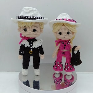 May include: Two crocheted dolls wearing cowboy hats and outfits. The doll on the left is wearing a black and white outfit with a white hat. The doll on the right is wearing a pink outfit with a white hat and a black purse.