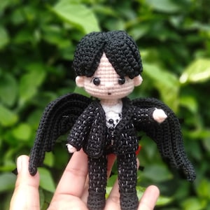 May include: A crocheted doll wearing a black suit with wings. The doll has black hair and a white face with black eyes.