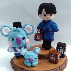 May include: A blue and white crocheted koala bear wearing a black hat and a blue and white patterned shirt. The koala is standing on a wooden surface with a blue crocheted person holding a lantern. There are also two brown crocheted lanterns on the surface.