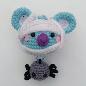 Könnte beinhalten: Ein gehäkeltes blaues und weißes Mumienmonster mit einer lila Nase und schwarzen Augen. Das Monster ist an eine graue gehäkelte Spinne mit schwarzen Beinen befestigt.
