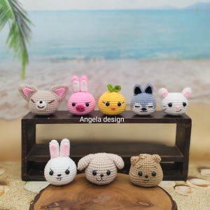 May include: A collection of seven crocheted animal heads, including a fox, bunny, chick, wolf, and mouse, displayed on a wooden shelf. The animals are in various colors, such as pink, yellow, and gray. The text "Angela design" is visible.