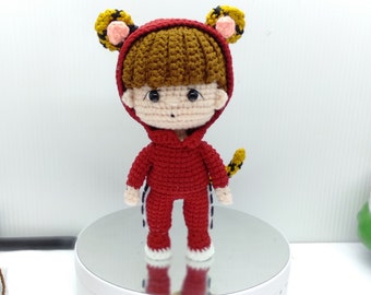 May include: A crocheted tiger costume for a small doll. The costume is red with black stripes and yellow ears. The doll has brown hair and a pink face.