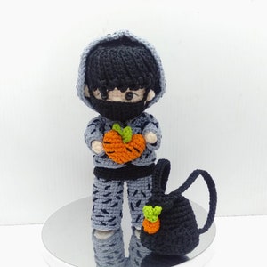 Puede incluir: Una muñeca de crochet que lleva un suéter con capucha gris, pantalones negros y una máscara facial negra. La muñeca sostiene una zanahoria de crochet naranja y tiene una mochila de crochet negra con una zanahoria verde en ella.