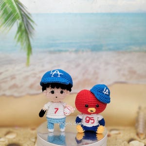 May include: Two crocheted dolls, one a boy and the other a bear, both wearing blue baseball caps with the letter "A". The boy doll holds a baseball and wears a jersey with the number "7". The bear doll wears a jersey with the number "95".