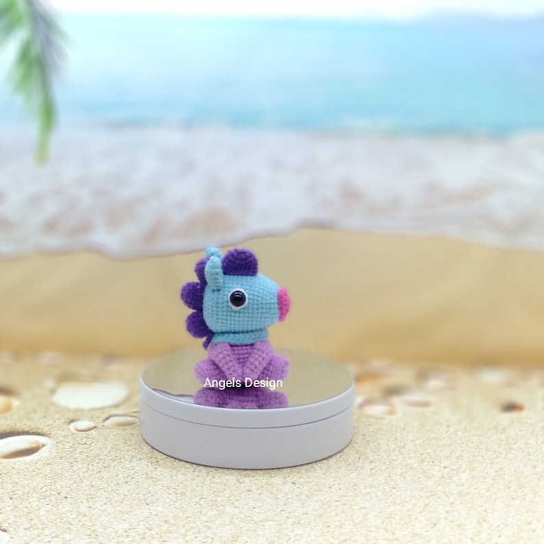K&ouml;nnte beinhalten: Ein geh&auml;keltes blaues und violettes Pferdespielzeug mit einer rosa Nase und einem schwarzen Auge. Das Spielzeug sitzt auf einer wei&szlig;en runden Plattform mit dem Text "Angels Design" darauf. Der Hintergrund ist ein verschwommenes Bild eines Strandes.