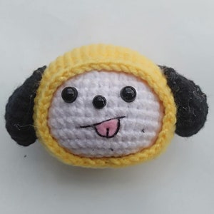 May include: A crocheted plush toy dog with yellow and black fur, wearing a yellow hat. The dog has white fur on its face with black eyes and a pink tongue sticking out.