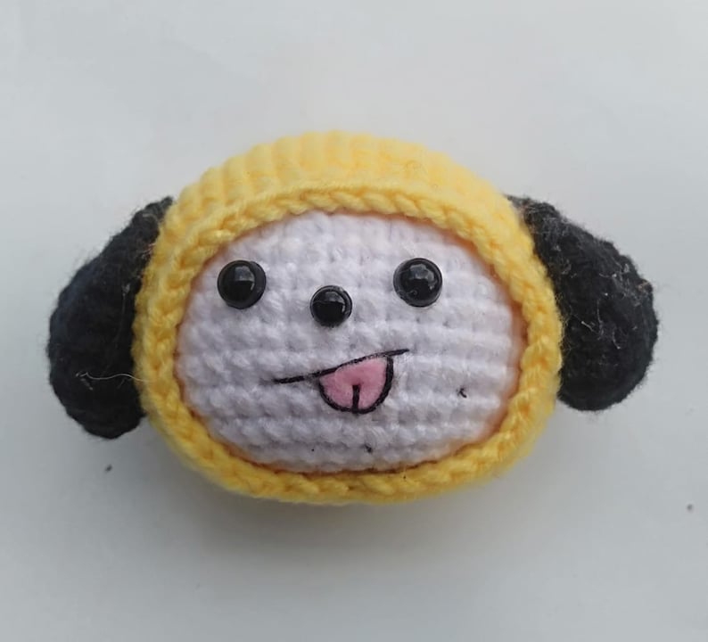 May include: A crocheted plush toy dog with yellow and black fur, wearing a yellow hat. The dog has white fur on its face with black eyes and a pink tongue sticking out.