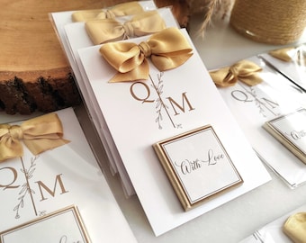Könnte beinhalten: Ein Satz weißer Karten mit goldenen Schleifen und einer kleinen quadratischen Schokoladentafel mit dem Text "With Love". Die Karten tragen die Initialen "QM" und ein florales Design.
