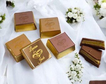 May include: An assortment of chocolate squares with gold foil wrapping. The tops of the chocolates are brown and feature the names "Lauren" and "Jack" with a ring design. Some chocolates are cut to reveal a layered filling. White flowers are scattered around.