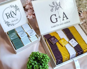 Könnte beinhalten: Zwei weiße Geschenkboxen gefüllt mit Schokoriegeln. Eine Box hat die Initialen "F&V" und das Datum "10.4.25" auf dem Deckel, die andere "G&A" und "GARETH AND AMBER". Die Pralinen sind in verschiedenen Farben, darunter Grün, Blau, Gelb und Braun.
