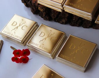 May include: Gold favor boxes with the initials "DC" and the names "Daniel & Carla" engraved on them. The boxes are arranged on a wooden surface.