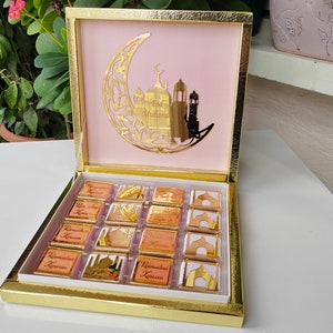 Könnte beinhalten: Eine goldene Geschenkbox mit einem rosafarbenen Deckel und einem goldenen Halbmond-Design. Der Deckel trägt den Text "Eid Mubarak". Die Box ist geöffnet und enthält 16 kleine Schokoladenquadrate mit goldenen Folienverpackungen. Jedes Quadrat trägt den Text "Ramadan Kareem".