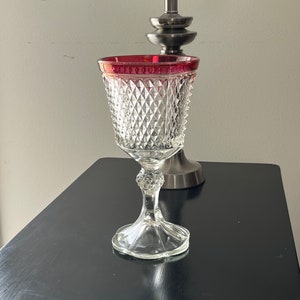 May include: A clear glass goblet with a diamond pattern and a red rim sits on a black surface. The goblet has a tall stem and a round base.
