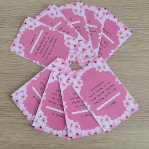 May include: Pink cards with a floral border and text. The cards are arranged in a fan shape. The text on the cards is in German. The cards have a pink background with white cherry blossoms.