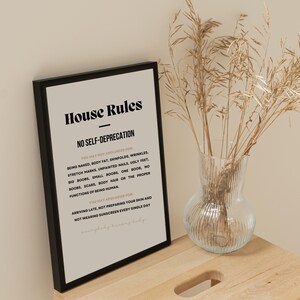 Op de afbeelding: Een ingelijste print met de tekst "House Rules" en "No Self-Deprecation" in zwarte letters. De print wordt weergegeven op een houten oppervlak naast een heldere glazen vaas met gedroogd siergras.