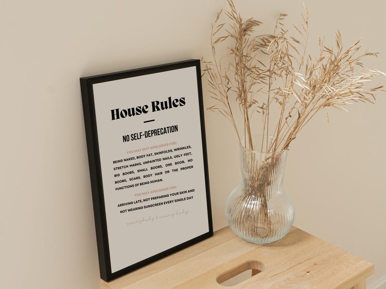 Op de afbeelding: Een ingelijste print met de tekst "House Rules" en "No Self-Deprecation" in zwarte letters. De print wordt weergegeven op een houten oppervlak naast een heldere glazen vaas met gedroogd siergras.