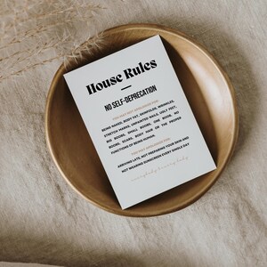 Op de afbeelding: Een witte kaart met de tekst "House Rules" en "No Self-Deprecation" in zwarte letters, geplaatst in een houten kom. De kaart bevat regels over lichaamsbeeld en zelfzorg. De kom staat op een beige linnen oppervlak.
