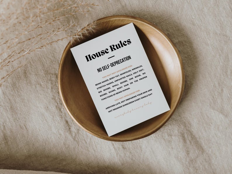 Op de afbeelding: Een witte kaart met de tekst "House Rules" en "No Self-Deprecation" in zwarte letters, geplaatst in een houten kom. De kaart bevat regels over lichaamsbeeld en zelfzorg. De kom staat op een beige linnen oppervlak.