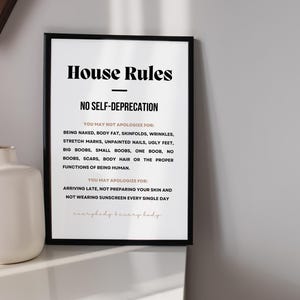 Op de afbeelding: Een ingelijste print met de tekst "House Rules" en "No Self-Deprecation". De tekst somt dingen op waarvoor je je niet hoeft te verontschuldigen, inclusief lichaamskenmerken, en dingen waarvoor je je wel moet verontschuldigen. Een witte vaas staat links.