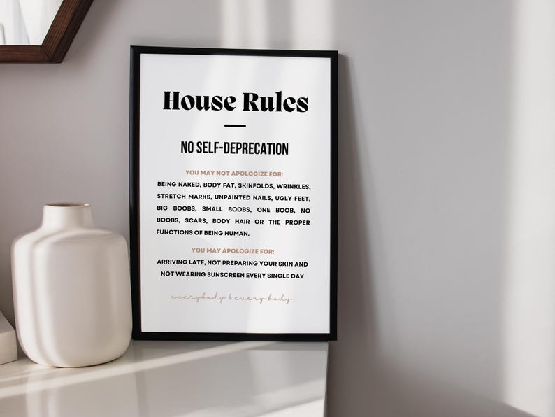 Op de afbeelding: Een ingelijste print met de tekst "House Rules" en "No Self-Deprecation". De tekst somt dingen op waarvoor je je niet hoeft te verontschuldigen, inclusief lichaamskenmerken, en dingen waarvoor je je wel moet verontschuldigen. Een witte vaas staat links.