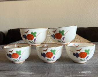 May include: Set of five white ceramic teacups with a floral design. The cups feature red, orange, green, and purple flowers with gold accents.