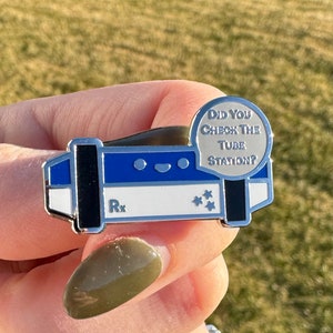 May include: A silver enamel pin shaped like a tube station with a blue and white design. The pin has a white background with the text "Rx" and a black background with five white stars. The pin also has a silver circle with the text "Did you check the tube station?" in black text.