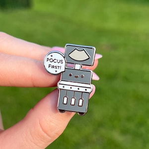 May include: Silver enamel pin featuring a cartoon ultrasound machine with a heart monitor and the text "POCUS FIRST!" in a speech bubble.
