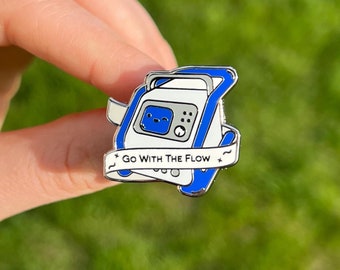 May include: A white and blue enamel pin with a cartoon illustration of a medical pump. The pin has a blue ribbon with the text "Go With The Flow" in white.