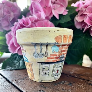 Puede incluir: Una maceta de terracota pintada a mano con una escena de cocina. La maceta presenta una pared de ladrillo, una estufa y utensilios de cocina.