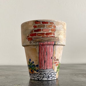 May include: A terracotta flower pot painted with a whimsical scene of a small house with a red door and a brick facade. The pot is decorated with small blue flowers and a stone pathway.