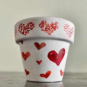 May include: White terracotta flower pot with red heart designs. The pot has a large heart in the center and smaller hearts scattered around the sides.
