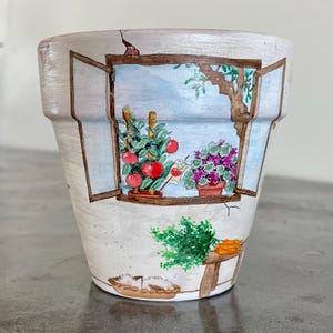 Könnte beinhalten: Ein handbemalter Terrakotta-Blumentopf mit einer Fensterszene. Das Fenster zeigt einen Blick auf einen Garten mit Tomatenpflanzen, Blumen und einem kleinen Tisch mit Karotten. Der Topf ist mit einer abgewetzten weißen Lackierung verziert.