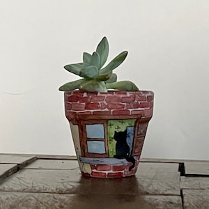 May include: A small terracotta planter with a painted brick design and a window with a black cat looking out. The planter is filled with a green succulent plant.