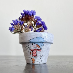Puede incluir: Un pequeño macetero de cerámica gris con un diseño pintado a mano de un gato con un sombrero azul y una bufanda roja y blanca. El macetero está lleno de flores moradas.
