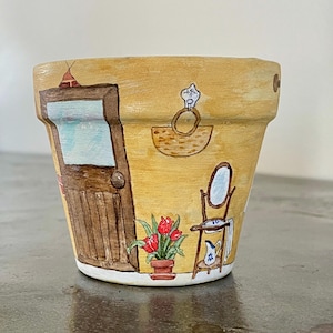 May include: A hand-painted terracotta flower pot with a yellow background and a scene of a house with a door, a mirror, a basket, and a potted plant with red flowers.