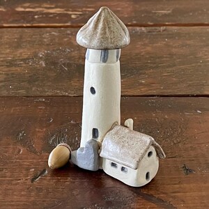 May include: A ceramic lighthouse figurine with a brown roof and a white base. The lighthouse is next to a smaller ceramic house with a brown roof and white walls. There is a small brown acorn at the base of the lighthouse.