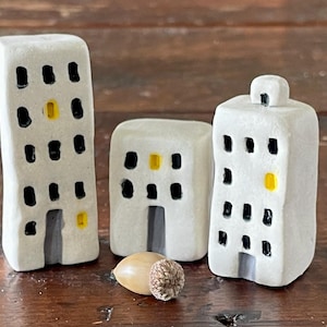 Op de afbeelding: Drie witte keramische gebouwbeeldjes met zwarte ramen en gele accenten. De gebouwen hebben grijze deuropeningen. Een eikel staat voor de gebouwen. De beeldjes staan op een bruin houten oppervlak.
