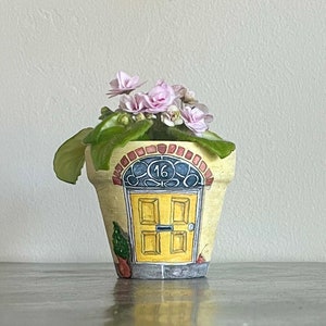 May include: A yellow ceramic planter with a painted door design and the number 16. The planter has a brown brick archway and a small green plant with pink flowers.